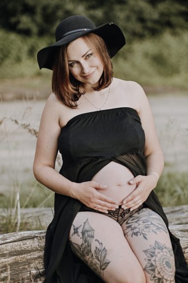 Schwangere Frau in einem schwarzen Kleid und Hut, lächelt und hält ihren Bauch.