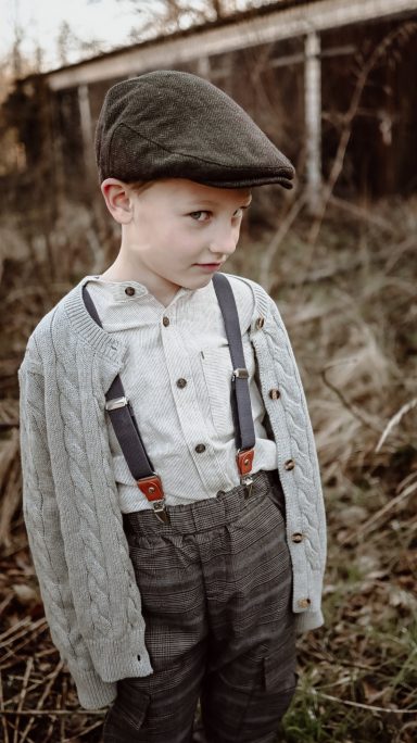 Ein Junge in einem grauen Cardigan und Schiebermütze steht in einem Feld.