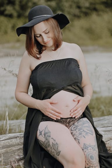 Schwangere Frau in Schwarz mit Hut, die ihren Bauch hält, umgeben von Natur.