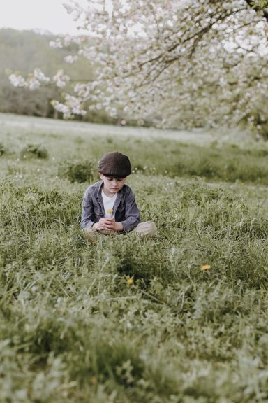 Ein Junge sitzt im Gras und betrachtet seine Hände, umgeben von blühenden Bäumen.