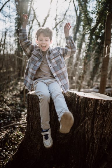 Junge sitzt fröhlich auf einem Baumstumpf im Wald und hebt die Arme in die Luft.