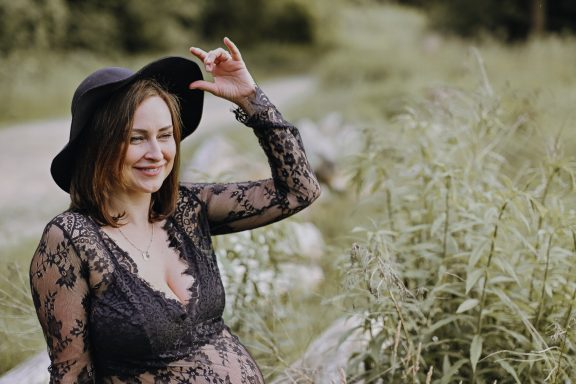 Schwangere Frau in schwarzem Hut und Spitzenkleid, lächelt im Freien.