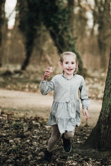 Ein Mädchen in einem grauen Kleid läuft fröhlich durch einen Wald mit Herbstlaub.