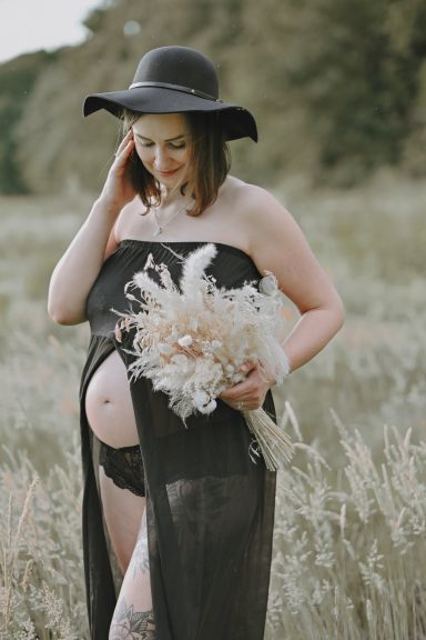 Schwangere Frau in schwarzem Kleid mit Hut und Trockenblumenstrauß in der Natur.