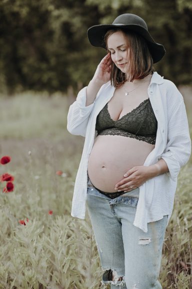 Schwangere Frau in Jeans und Hut steht in einem blühenden Feld mit roten Mohnblumen.