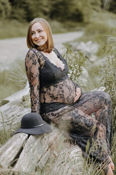 Schwangere Frau in schwarzer Spitzenkleidung, sitzt auf einem Baumstamm, mit Hut daneben.