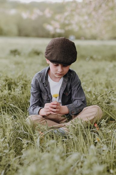 Ein Junge mit einer Mütze sitzt im Gras und hält eine Blume in der Hand.