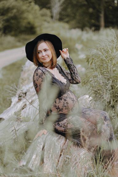 Frau in schwarzem Kleid und Hut sitzt auf einem Baumstamm in der Natur.